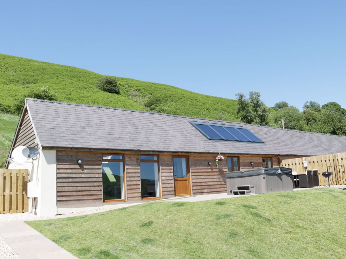 Timber-clad building at 1 Beacon View Barn with a lawn and hot tub below a green hillside
