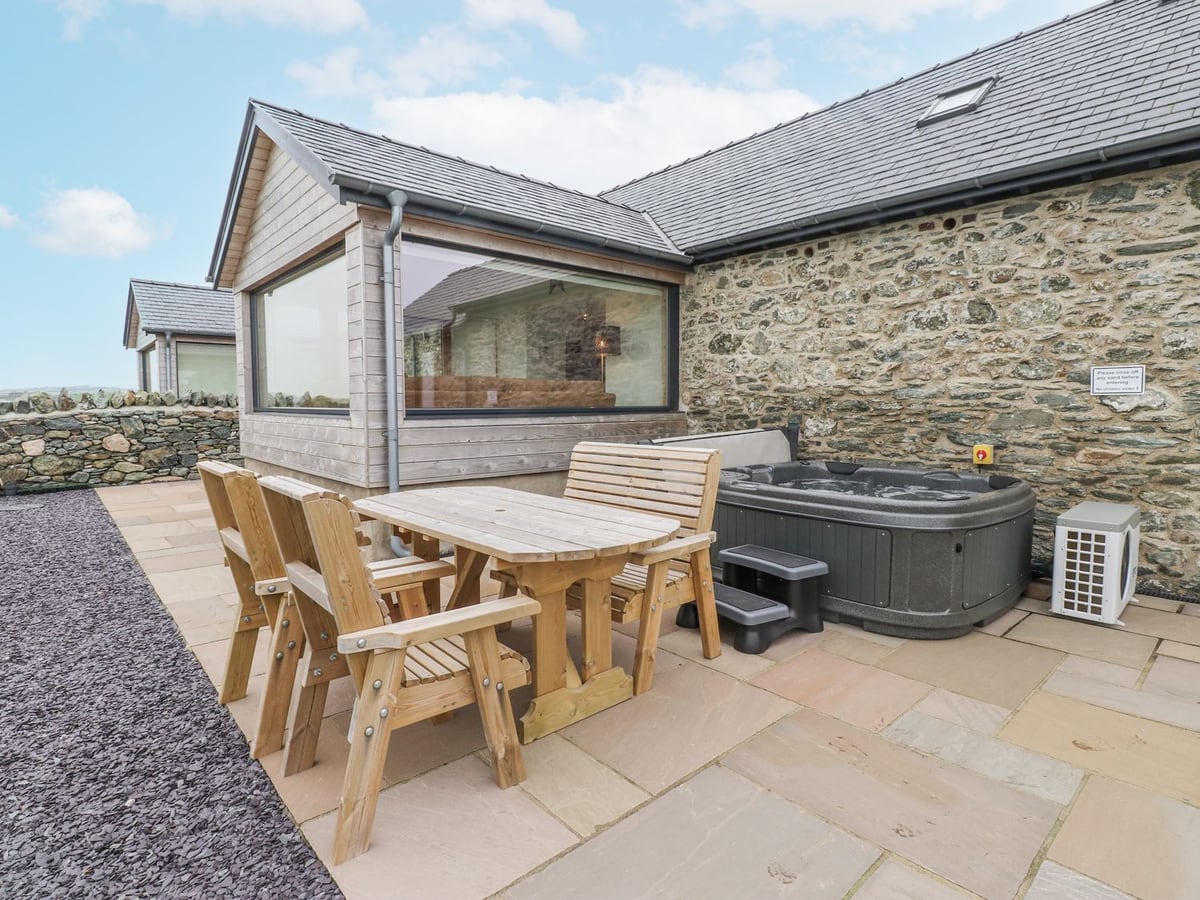 Paved patio at 2 Trem Y Mor with a wooden table and chairs beside a hot tub against a stone wall