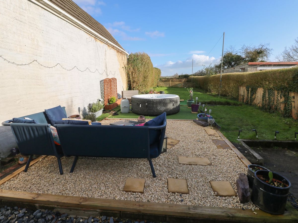 Gravel patio with blue sofa and an inflatable hot tub on a long lawn edged by hedge and fence