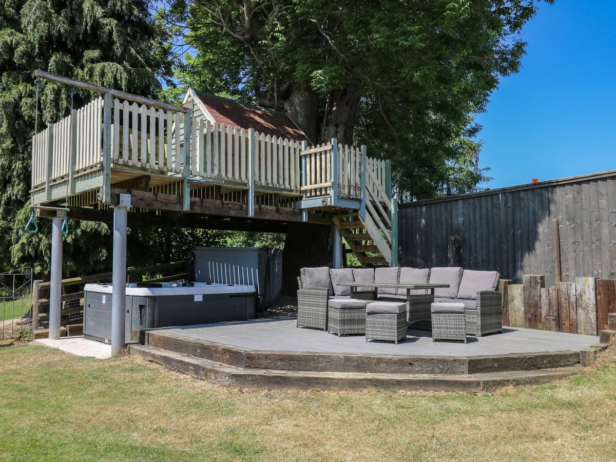 Awelfan decking with a hot tub and wicker sofa set beneath a tree house platform, beside a lawn