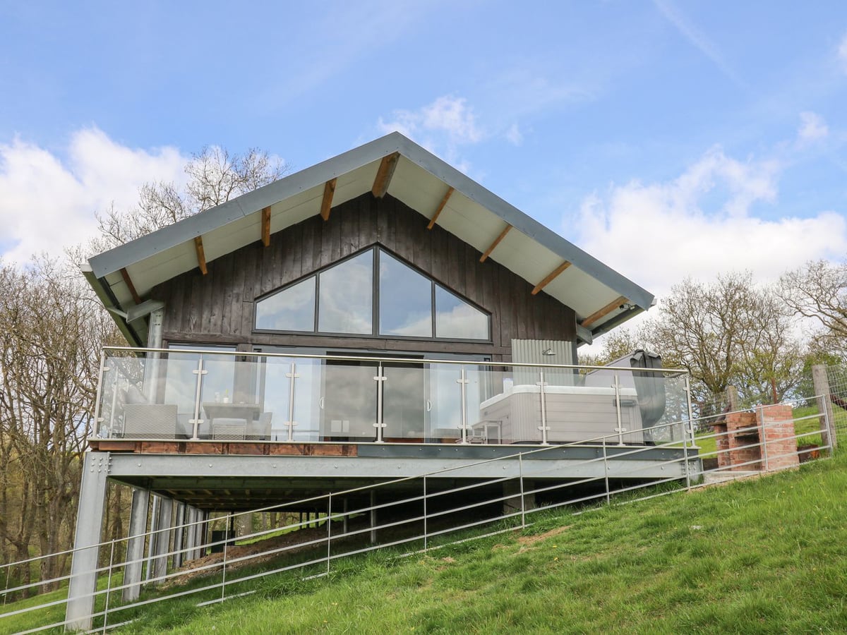 Barcud Coch lodge with a glass-railed raised terrace, hot tub and outdoor shower above a grassy slope