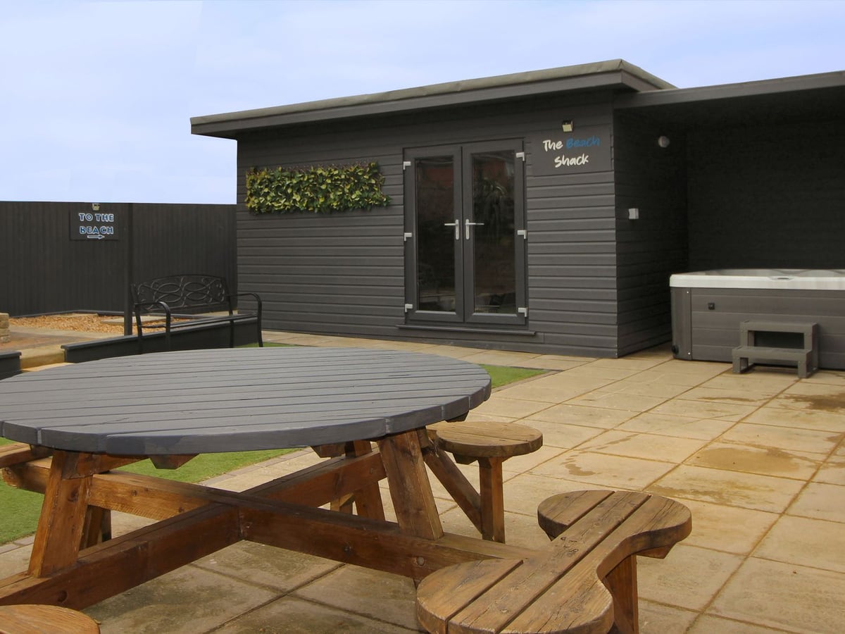 Paved rear patio at Beachway with a round picnic table, grey garden room and hot tub