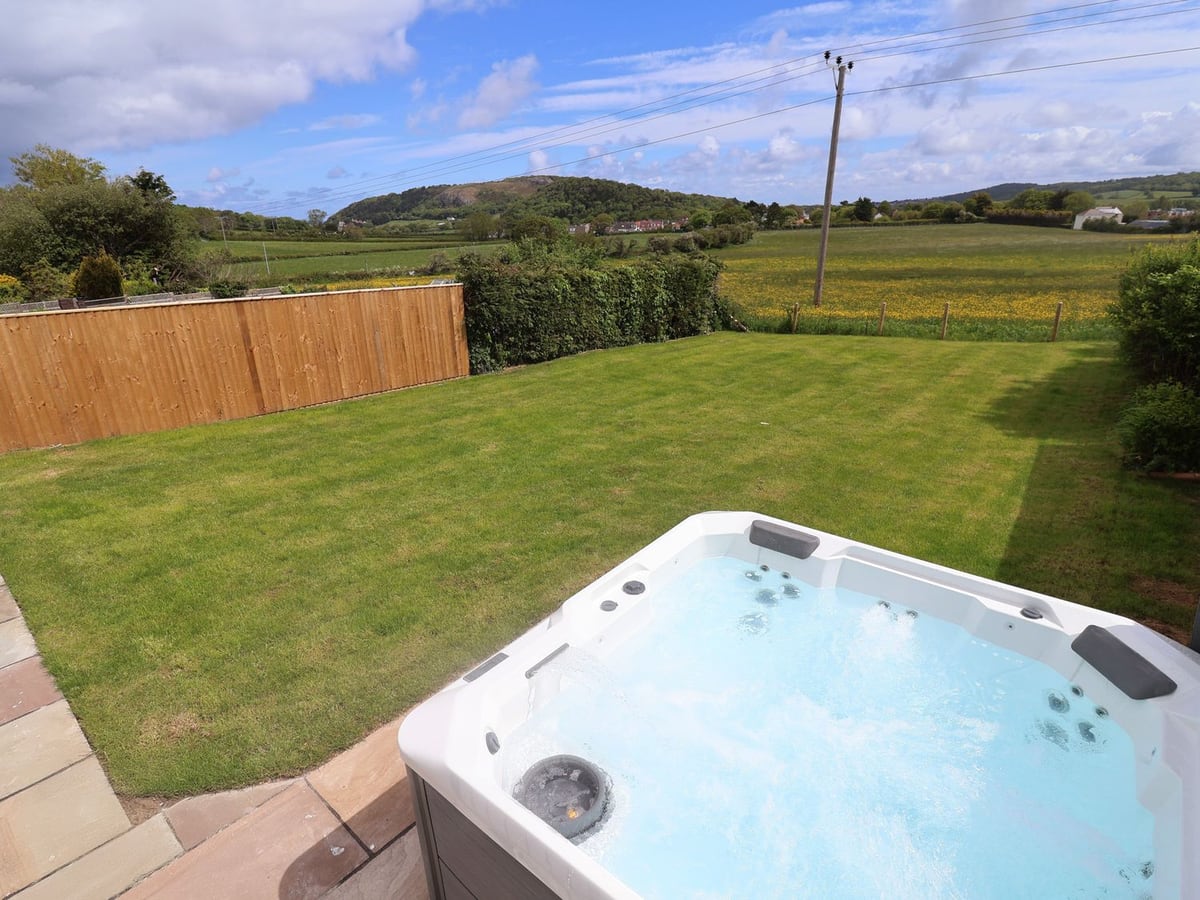 Bella Vista hot tub beside a lawn with a wooden fence and hedges, with fields and a hill beyond