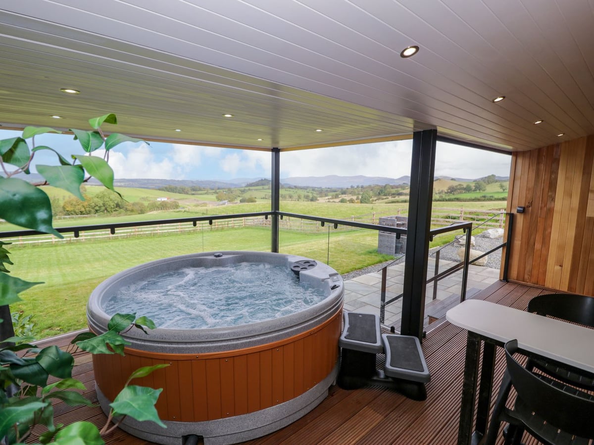 Hot tub on a roofed deck at Betsi (Lodge 1) with glass balustrade, looking over lawn and fields to hills