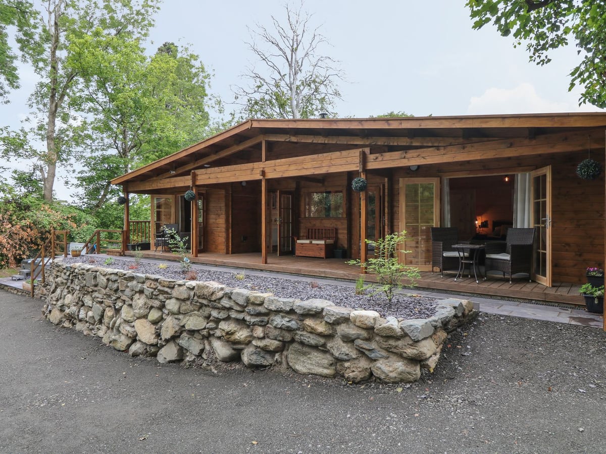 Bryn Derwen Lodge log cabin with a long roofed veranda, wicker chairs and a raised stone-walled bed