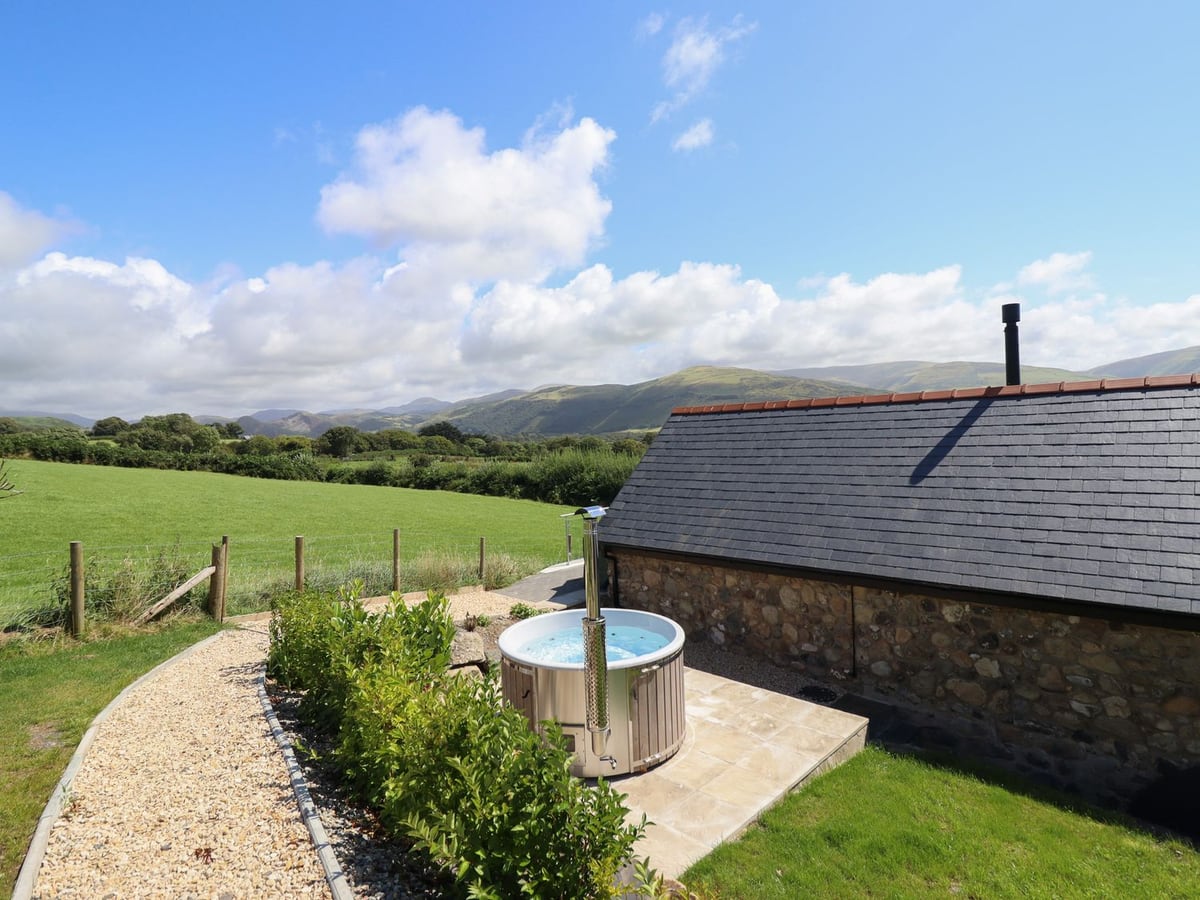 Bryn Eglwys Cottage wood-fired hot tub on a paved patio by a gravel path, lawn and hills