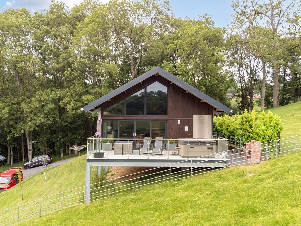 Bwncath lodge on a grassy slope with a raised glass-railed deck, loungers and hot tub with its lid on