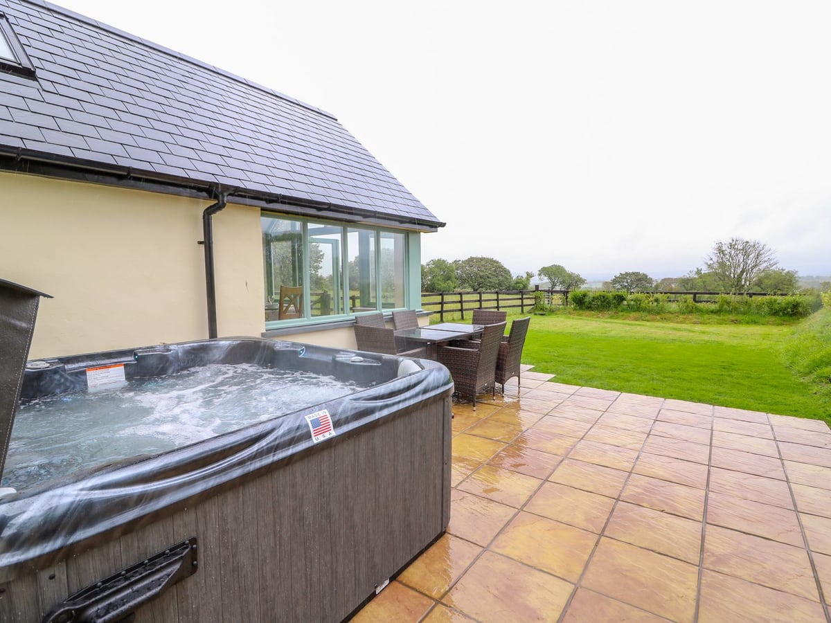 Open hot tub on a tiled patio at Bwthyn Eisteddfa Fach, with rattan seating and a fenced lawn beyond