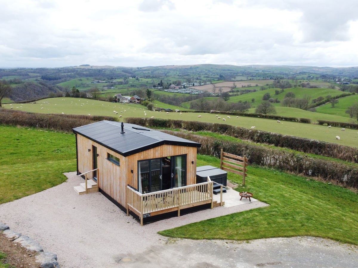 Aerial view of Cae Bedw cabin with a railed deck, hot tub with its lid on, lawn and rolling fields