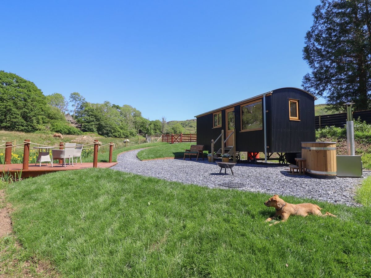 Celyn shepherd's hut with a dog lying on the lawn, wooden hot tub, fire pit and rope-railed deck