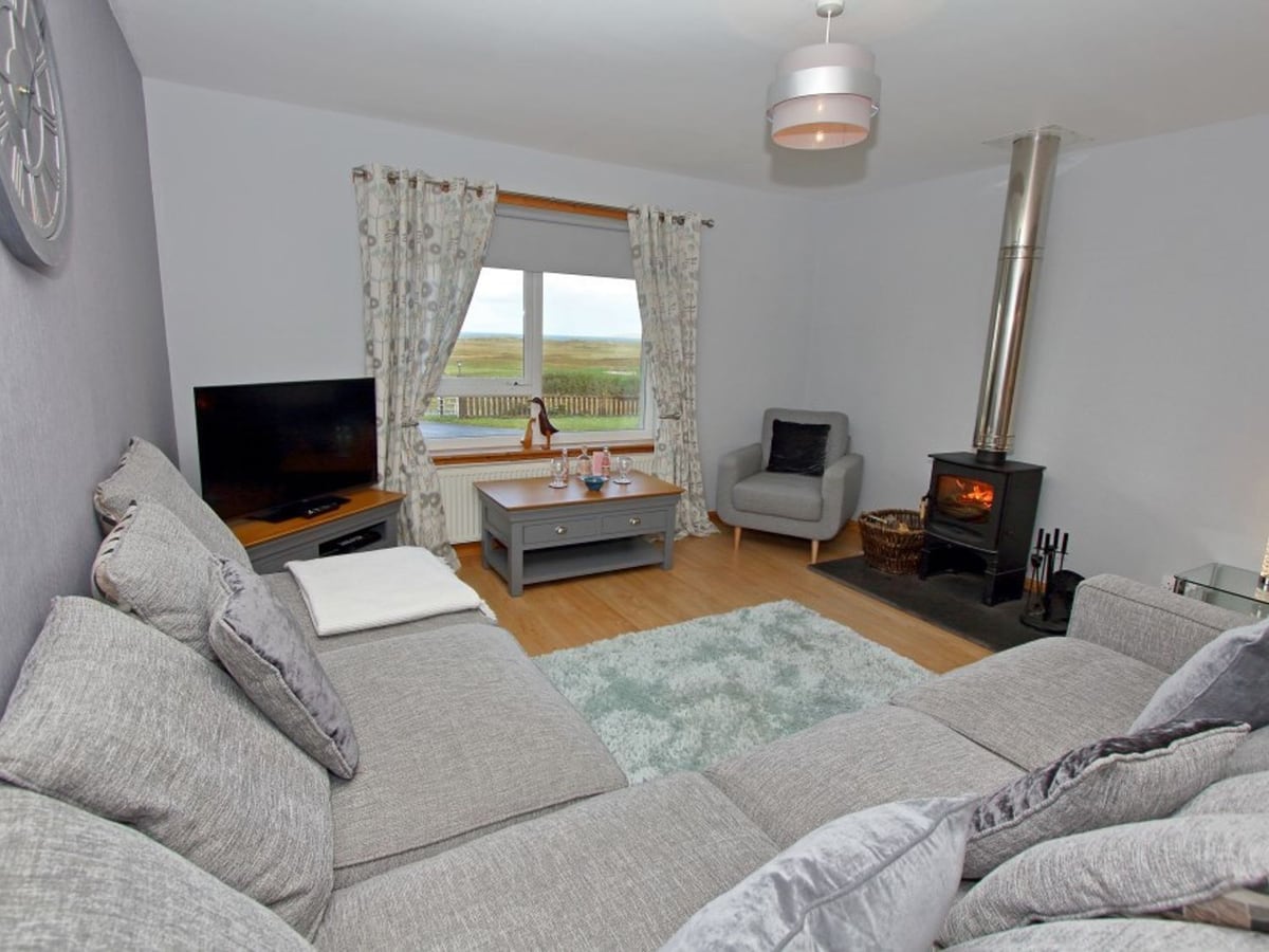 Clachan Sands Cottage living room with grey sofas, a stove and windows facing open grassland