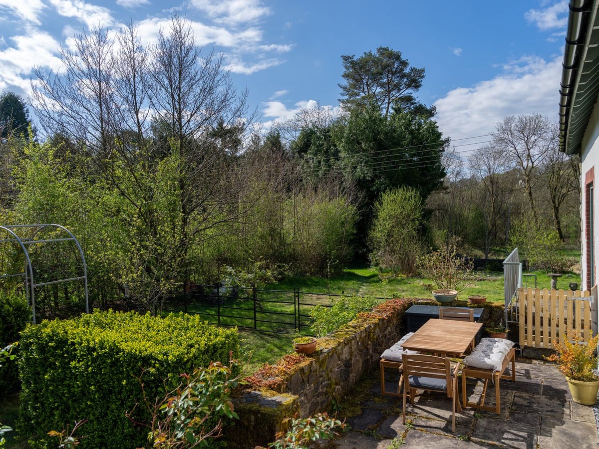 Stone-flagged patio with a wooden table beside Coach House, above a lawn with a metal railing and trees