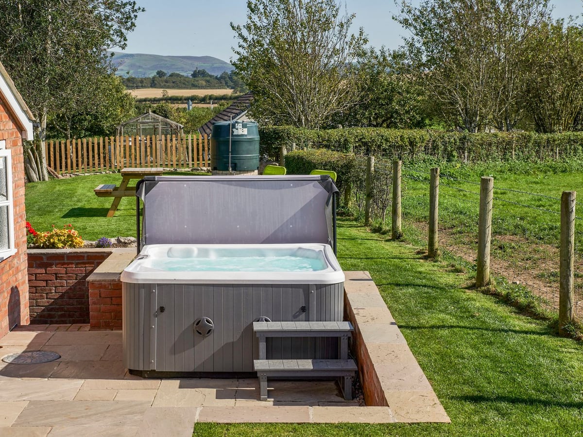 Conquer Hall hot tub on a paved terrace beside a lawn with picnic bench, fencing and distant hills