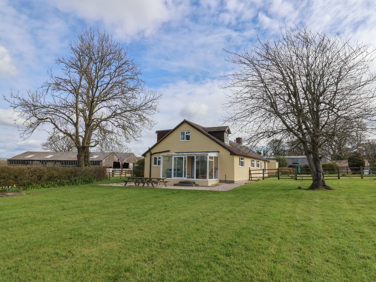Court House Farmhouse and its conservatory across a lawn with trees, fencing and farm buildings