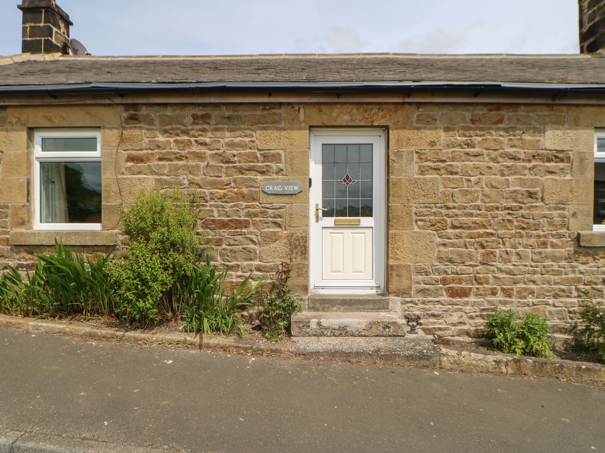 Stone frontage of Crag View with a white entrance door, cottage name plaque and planted border.