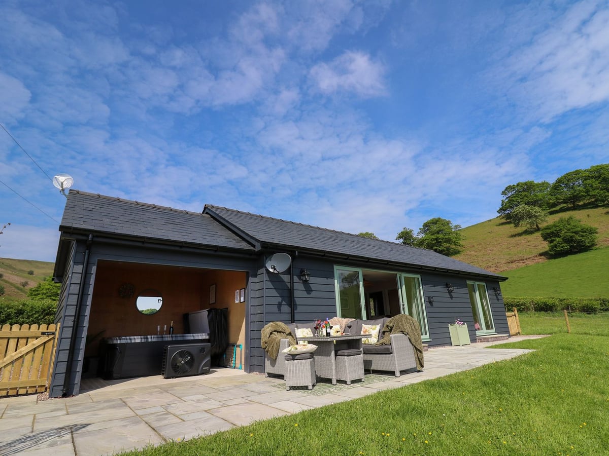 Dol Blodau dark clad lodge with patio seating, covered hot tub, lawn and wooden gate below green hills
