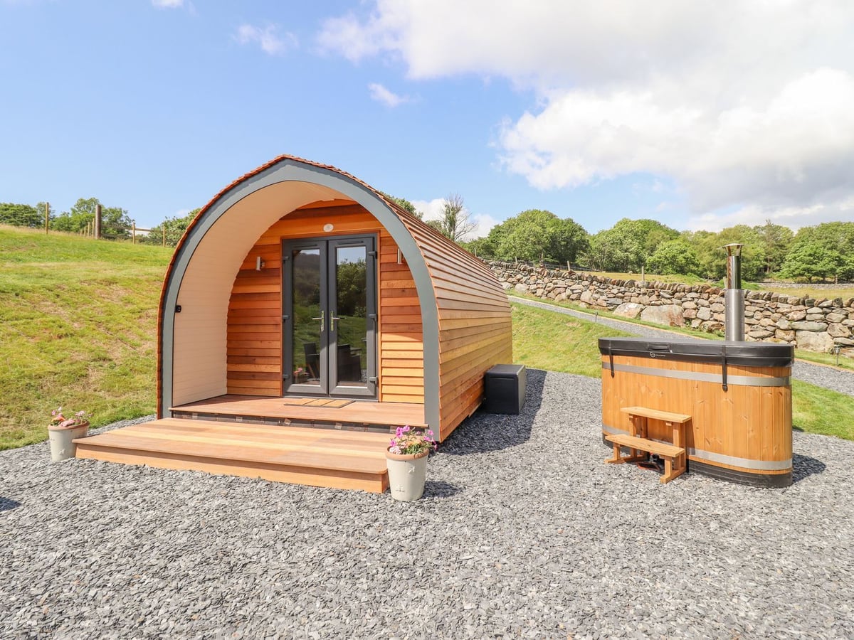 Eirlys pod exterior with private wooden hot tub on a gravel pitch beside grass
