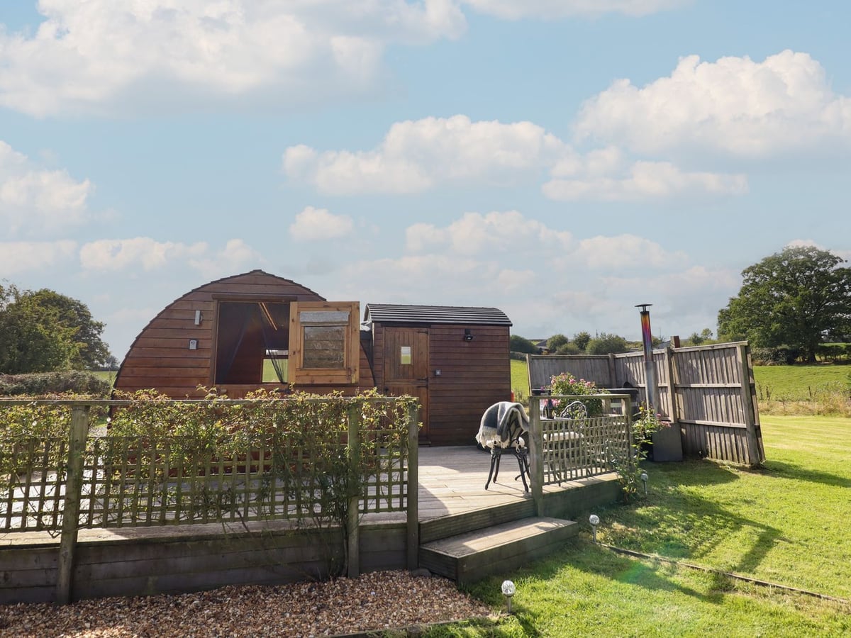 Embden Pod wooden glamping pod with trellis-edged deck, a chair and mown lawn beside fence panels