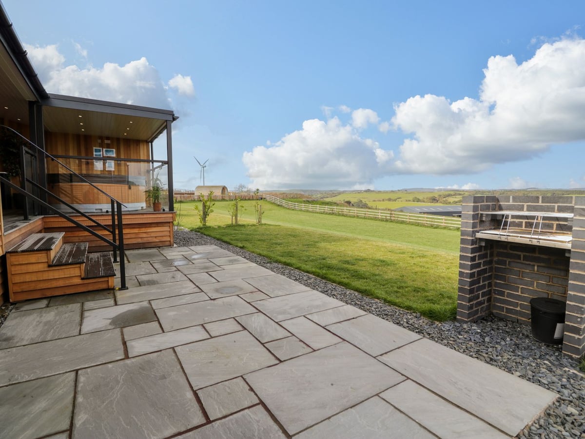 Enia lodge steps and covered deck beside a paved barbecue patio, open lawn and distant hills