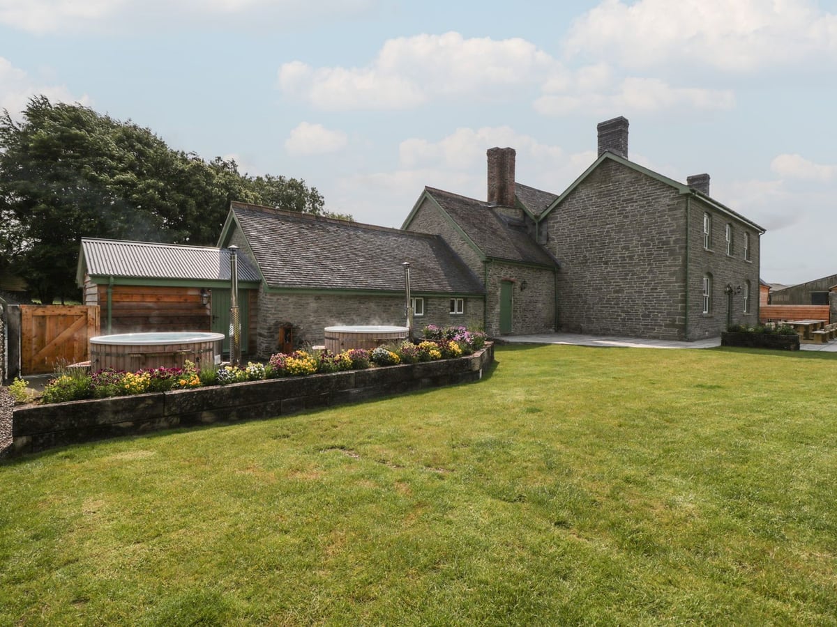 Evenjobb Court Farmhouse stone farmhouse with two wooden hot tubs behind a flower bed and a large lawn