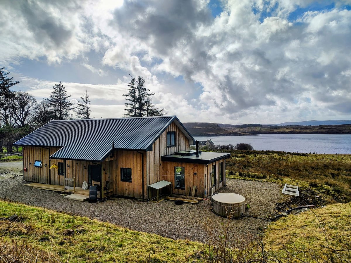 Faodail House timber exterior with a gravel terrace, round hot tub and a loch beyond open ground