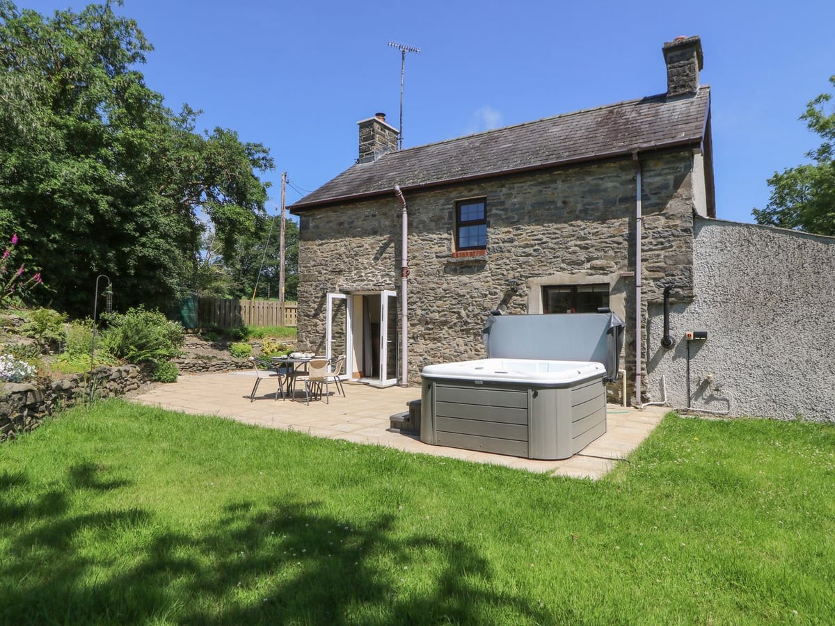 Stone cottage at Felin Gogoyan with a hot tub and patio table on paving beside a lawn and planted beds