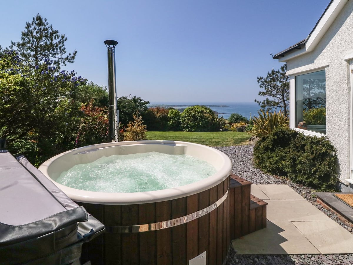 Wood-fired hot tub beside Fron Olau, with lawn, shrubs and a view to the sea and headland beyond