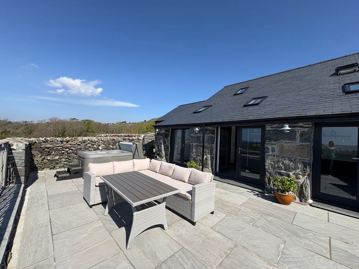 Paved patio at Gaerau Bach with a corner sofa, table, hot tub and dry stone walls by the stone cottage