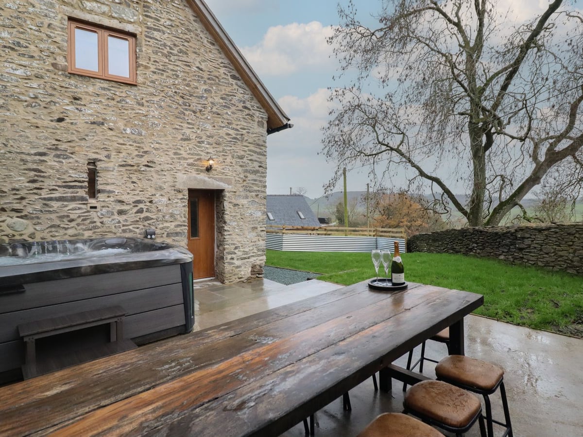 Hot tub and long outdoor table on a patio beside Godre'r Gyrn, with lawn and a stone wall