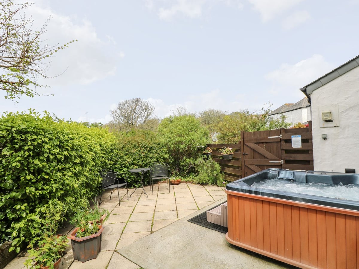 Hot tub beside Godrevy Barn's white exterior, with a paved patio, chairs, hedges and timber gate