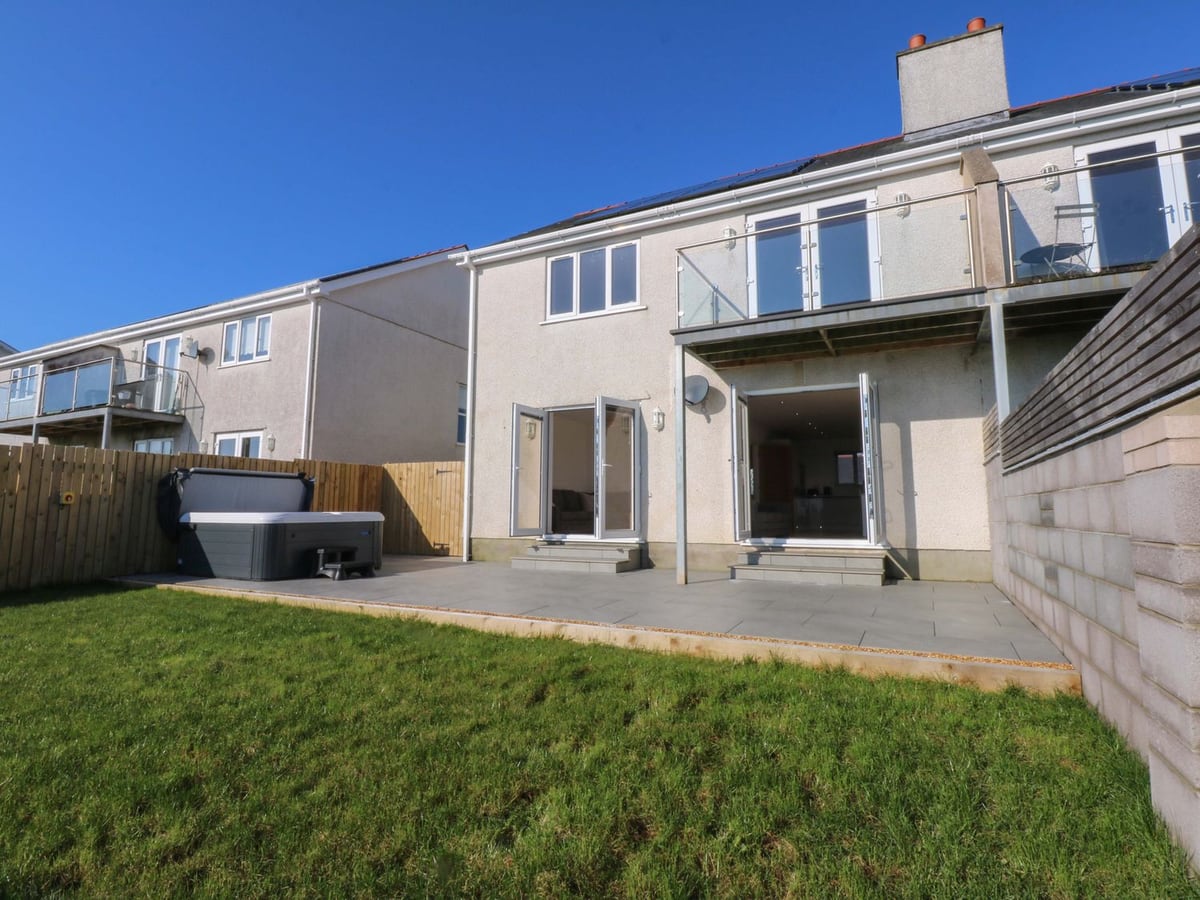 Golygfa rear lawn and paved patio with a hot tub, its lid raised, by a wooden fence and the house