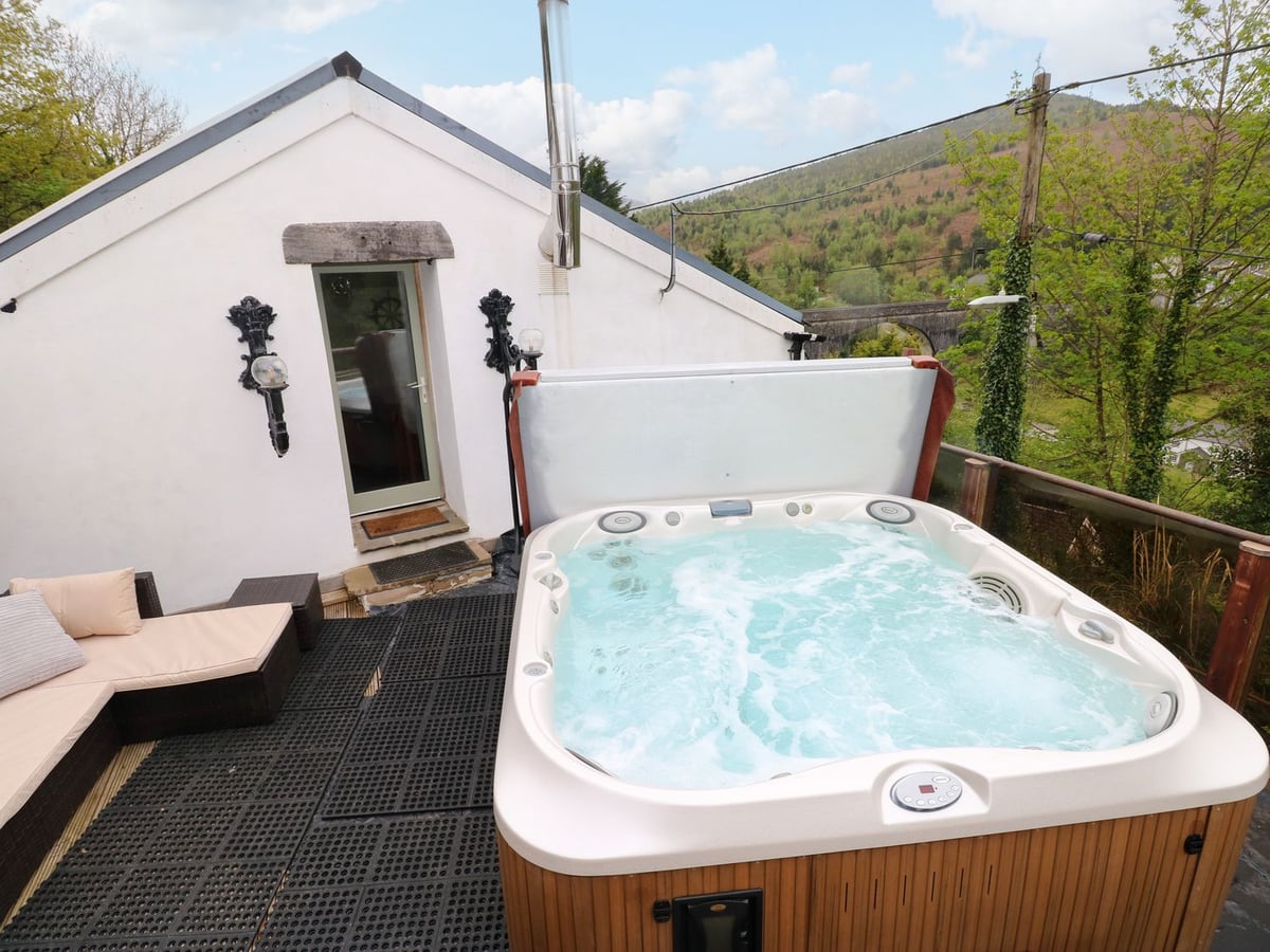 Glass-railed terrace at Greenheart Cottage with a sofa, hot tub with its lid raised and wooded hills