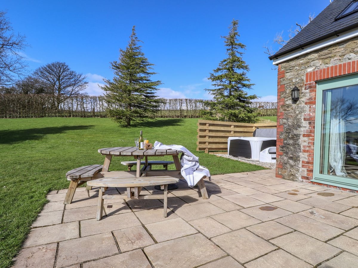 Paved patio with a picnic table beside Gwennol Cottage, a hot tub by a fence and open lawn