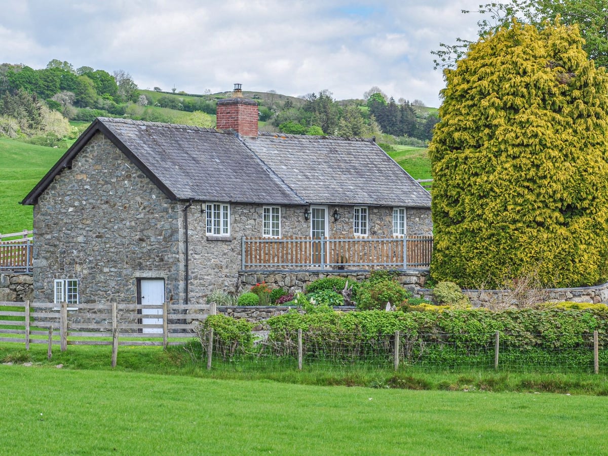 Hafan stone cottage with a fenced front terrace, planted beds and green hills beyond a field fence