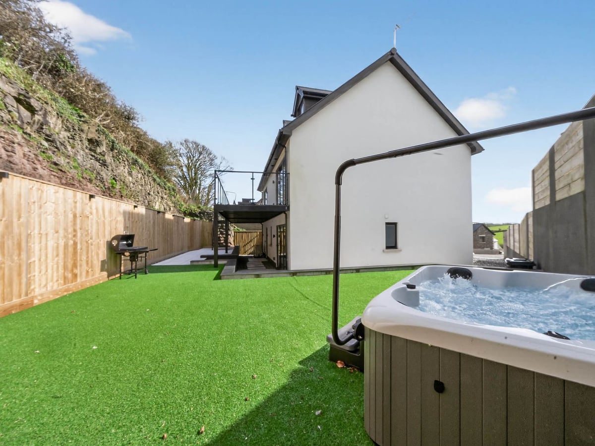 Heddfan hot tub on an artificial lawn with a barbecue, tall wooden fence and the white house beyond