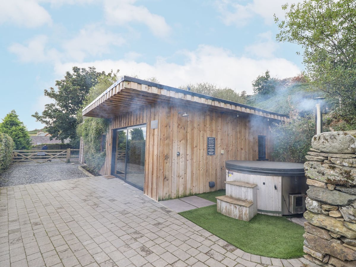 Hilltop Lodge, a timber-clad lodge with a wood-fired hot tub with its lid on, artificial turf and paving