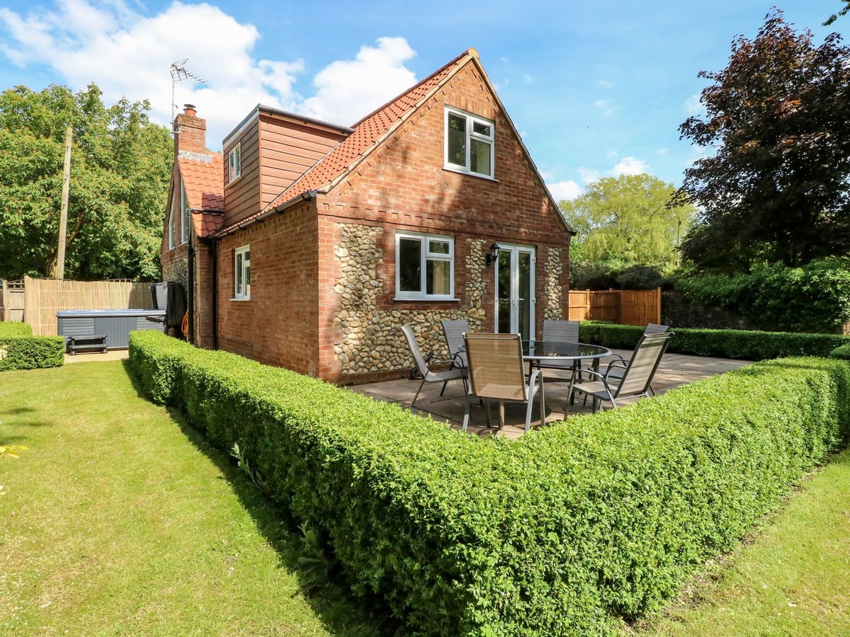 Keepers Cottage with brick and flint walls, patio seating, low hedges, lawn and a hot tub