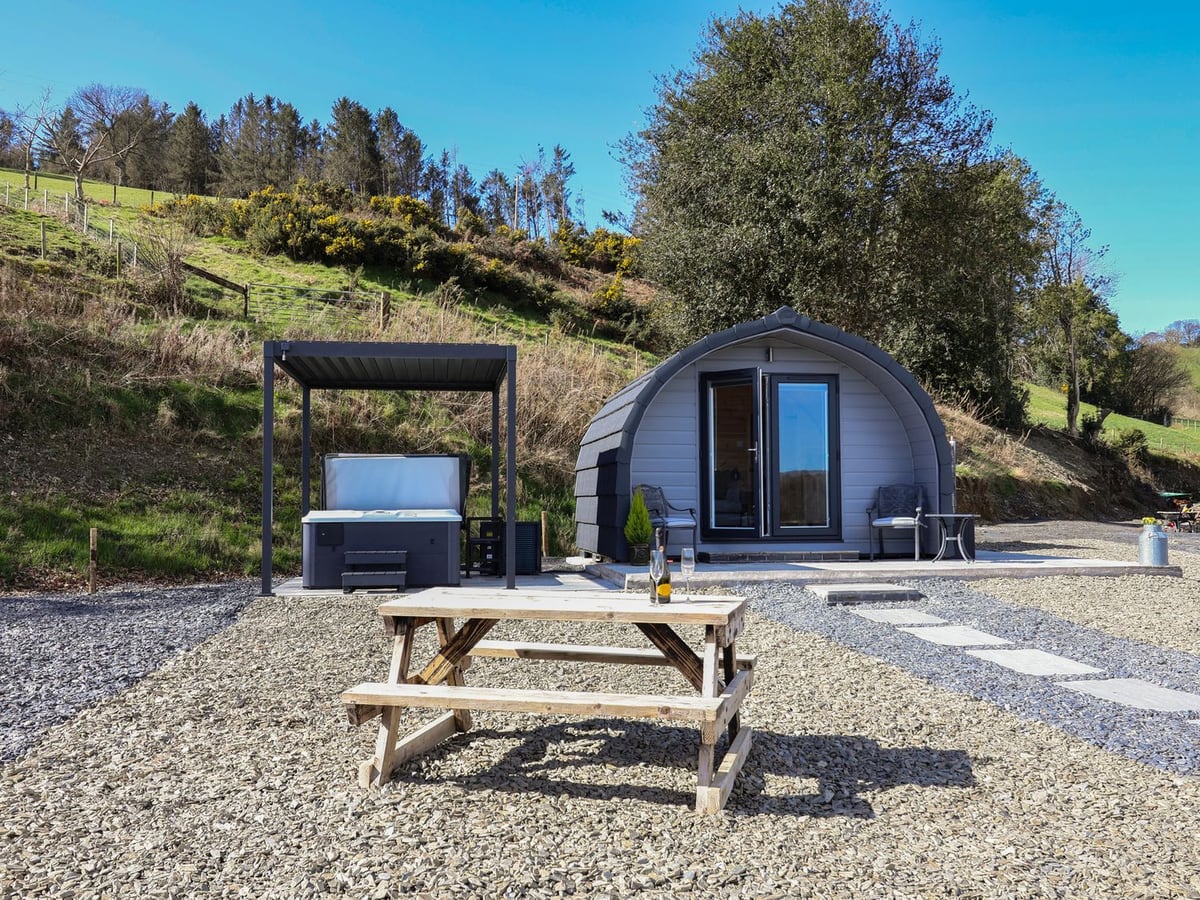 Lili Wen Fach grey pod with a hot tub under a canopy, a picnic bench on gravel and a wooded hillside