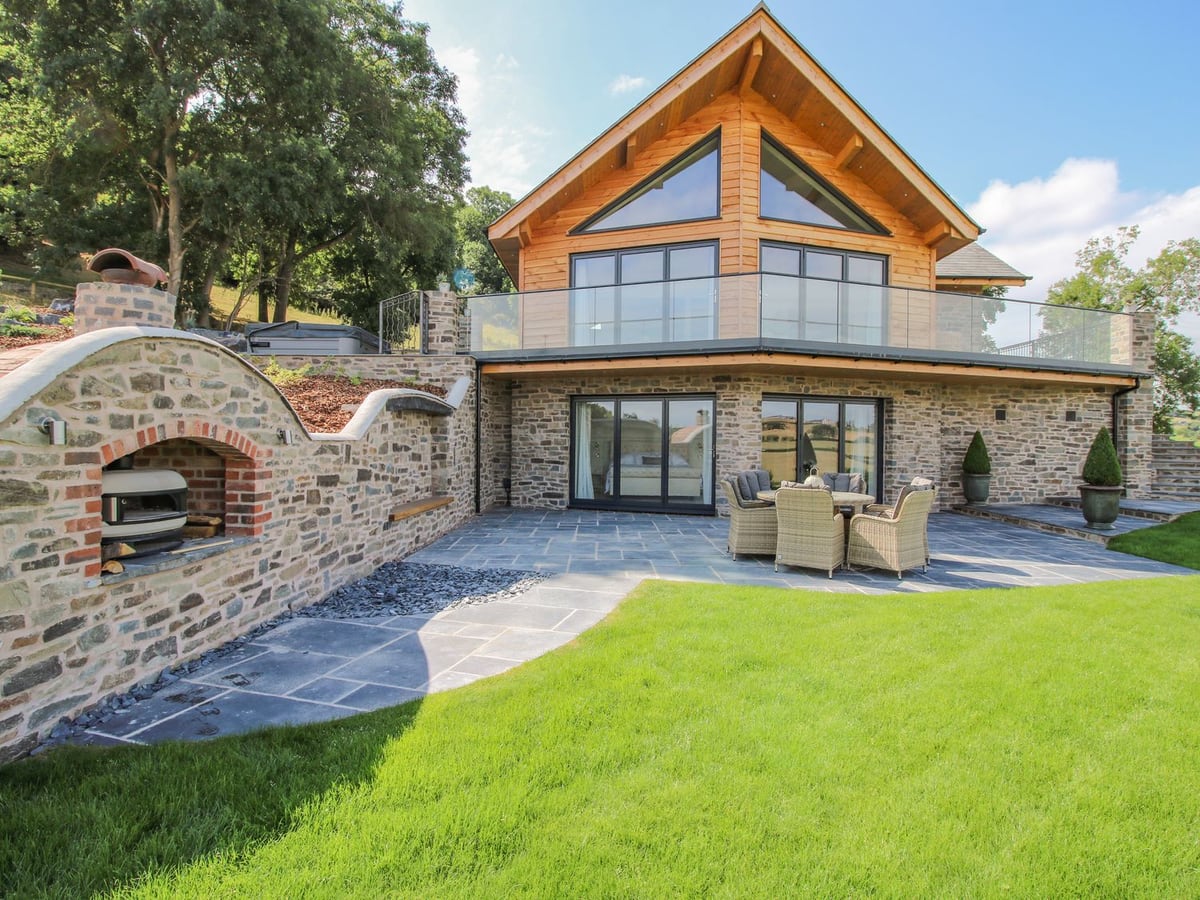 Llety Cariad lodge with a glass balcony above a slate patio with seating, a stone oven wall and lawn