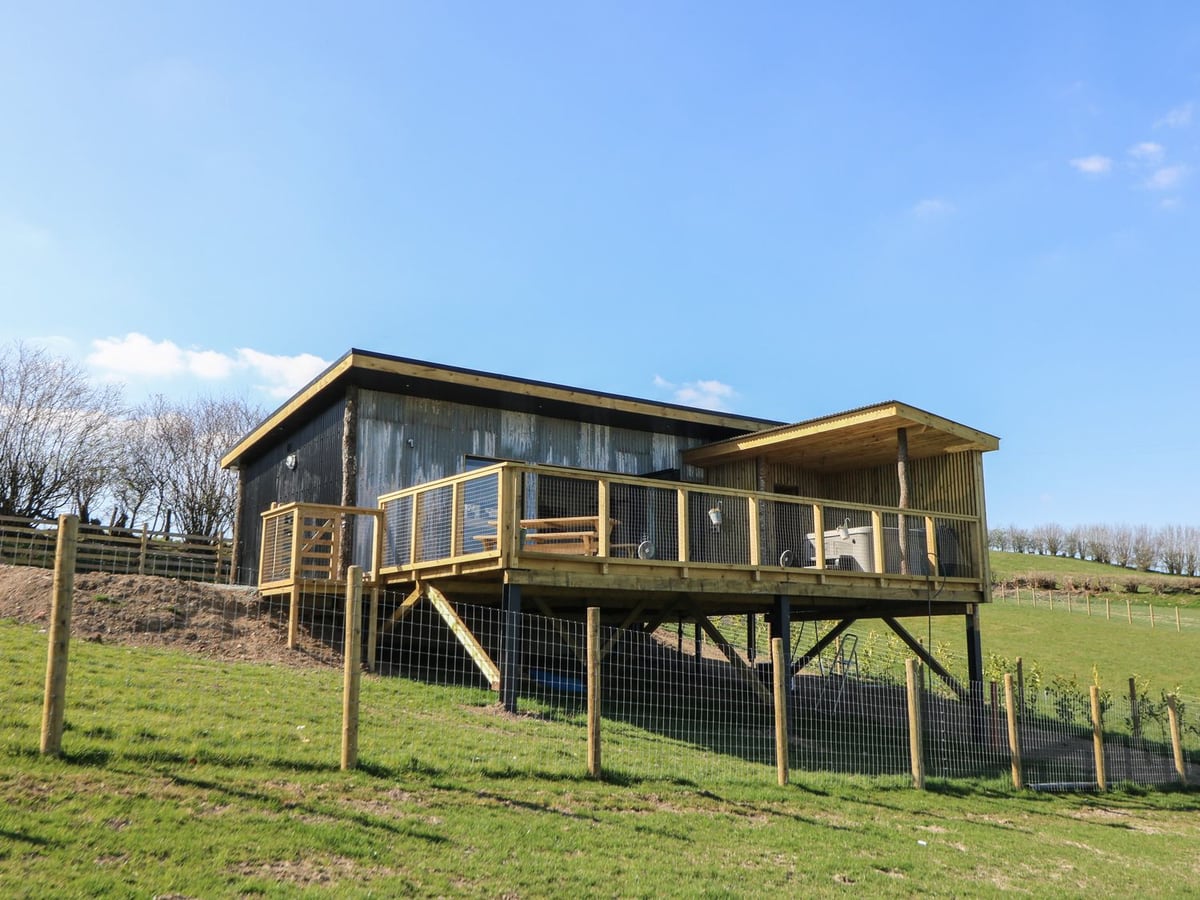 Llety Cariad timber lodge on a raised deck with hot tub and picnic table, above grass and a wire fence