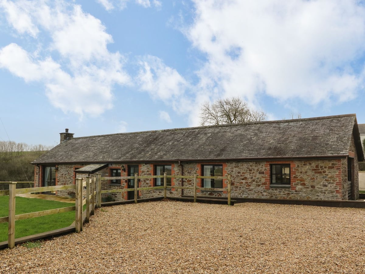 Long stone barn with glazed doors behind a timber gate and gravel approach at Long Linney