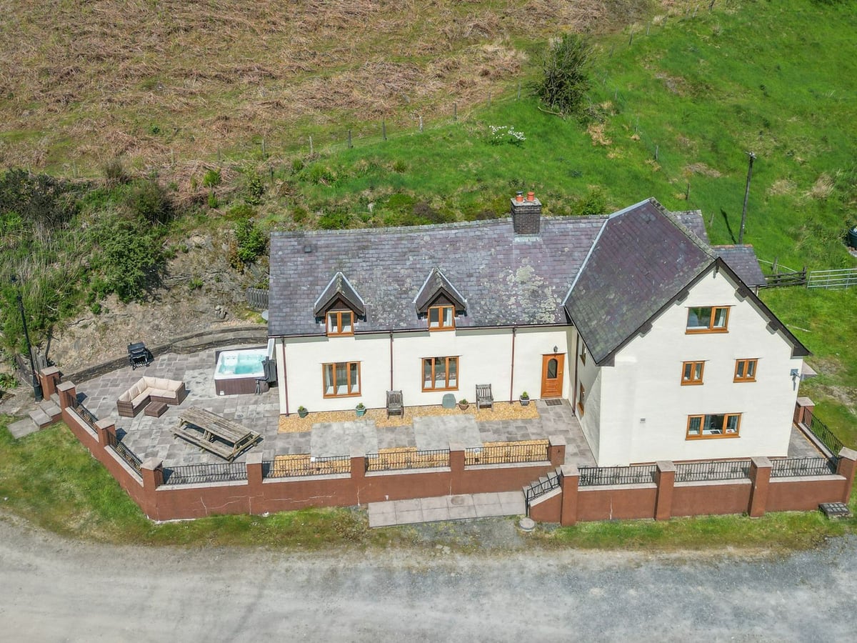 Aerial view of Maesgwyn farmhouse with a railed patio, hot tub, sofa and picnic bench below a hillside