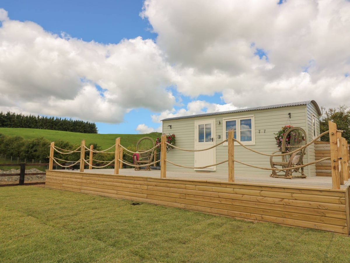 Marches Way shepherd's hut on a rope-railed timber deck above a lawn, with wicker chairs and green hills
