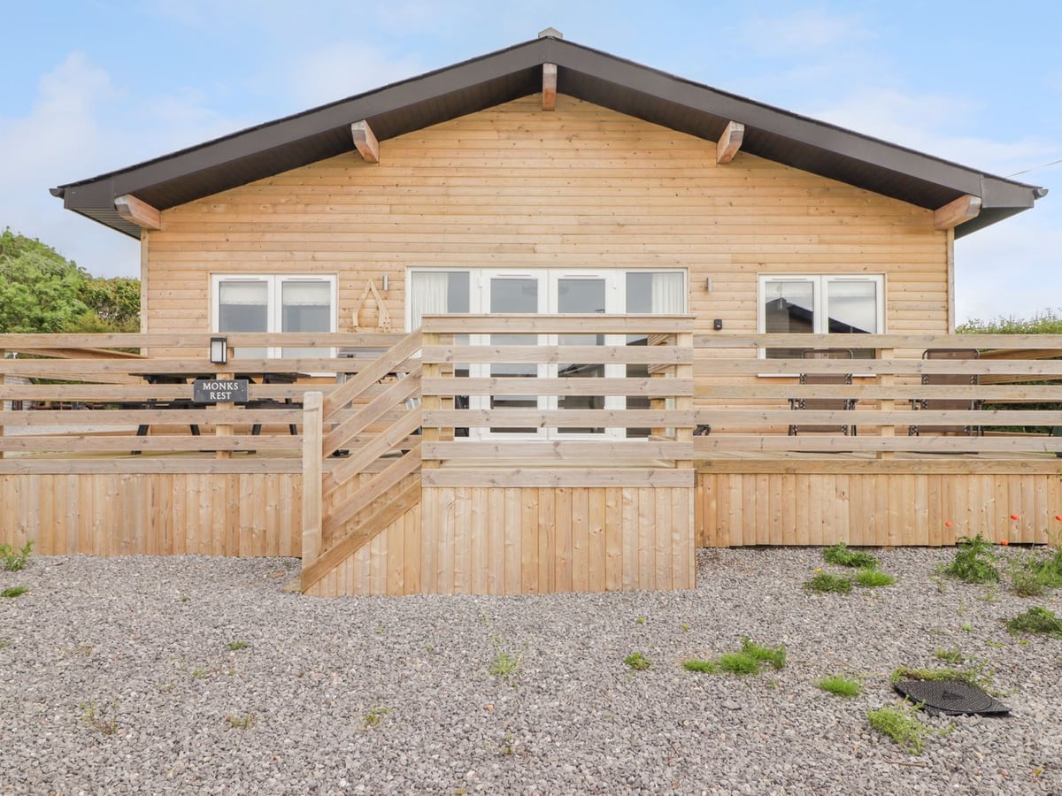 Monks Rest timber lodge front with white doors, railed decking and a named sign above gravel