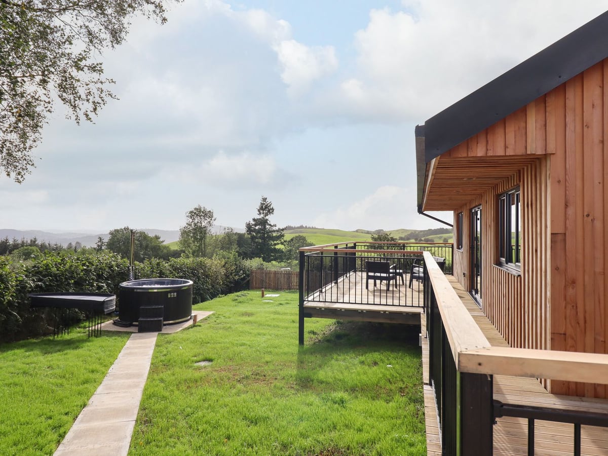 Mynydd Y Glyn Lodge lawn with round hot tub, hedge, railed deck with chairs and rolling hill views