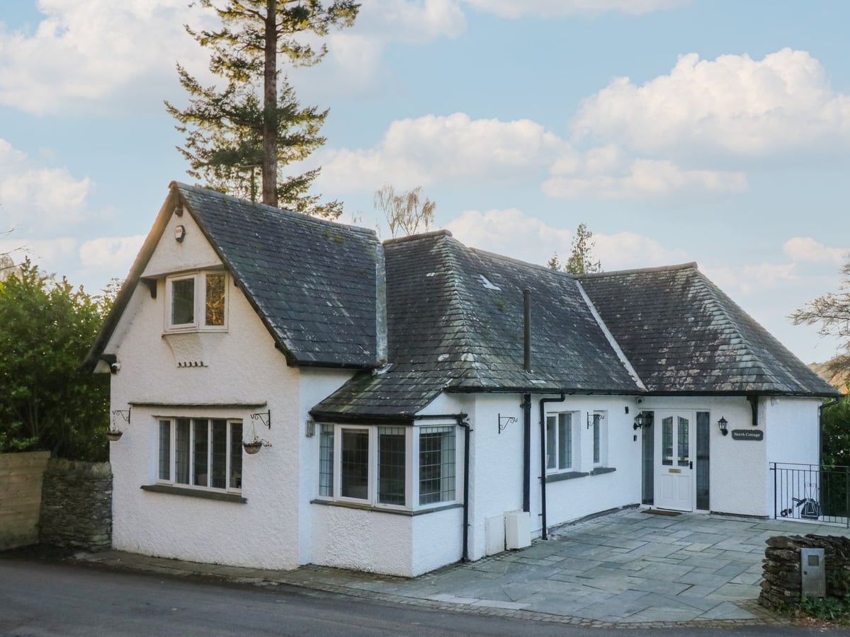 North Cottage white exterior with slate roofs, a bay window and a paved entrance beside the road