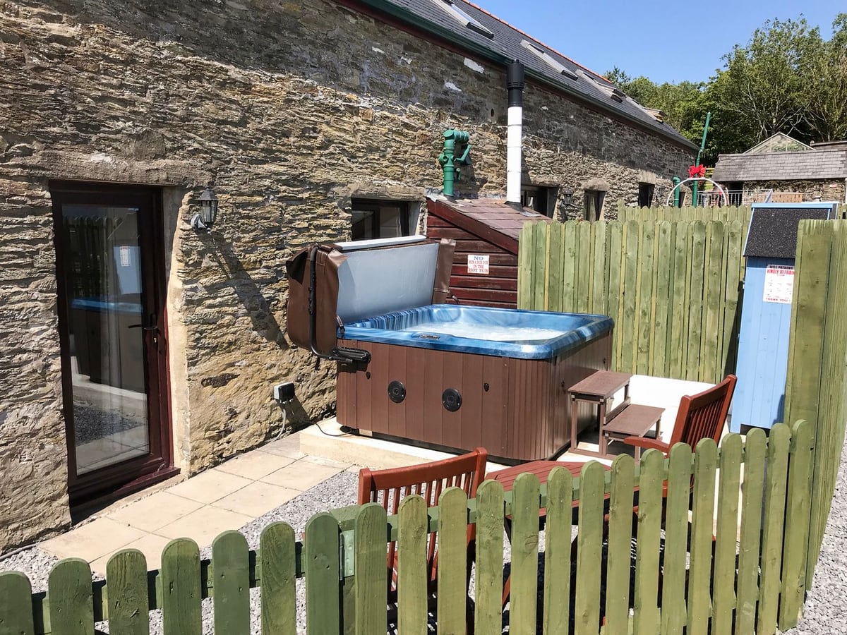 Hot tub on a paved patio beside Oak Cottage's stone wall, behind a green picket fence with chairs