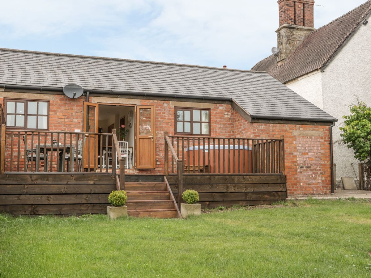 Old Hall Barn 1's brick exterior with a raised wooden deck, round hot tub and lawn