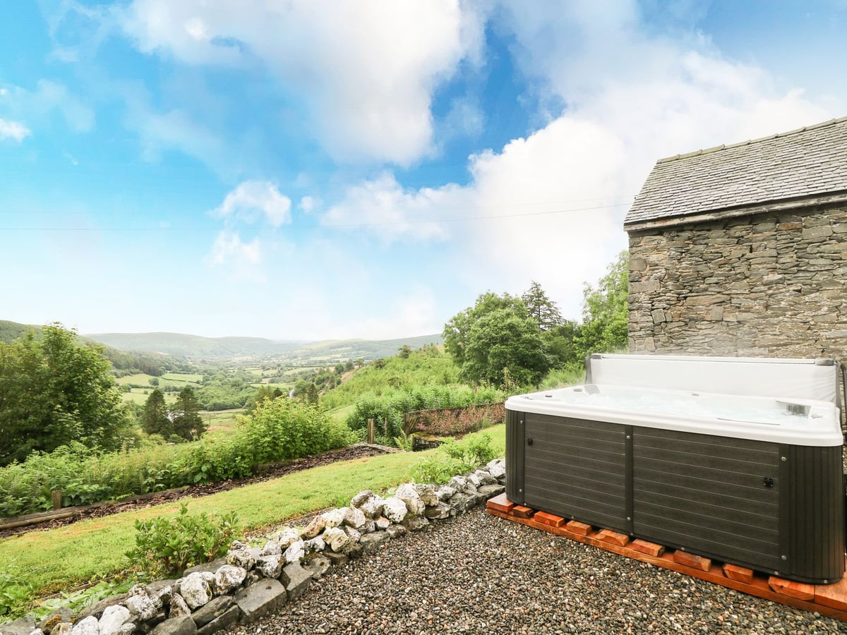 Hot tub with its lid raised on gravel beside a stone building, with a lawn and a green valley view