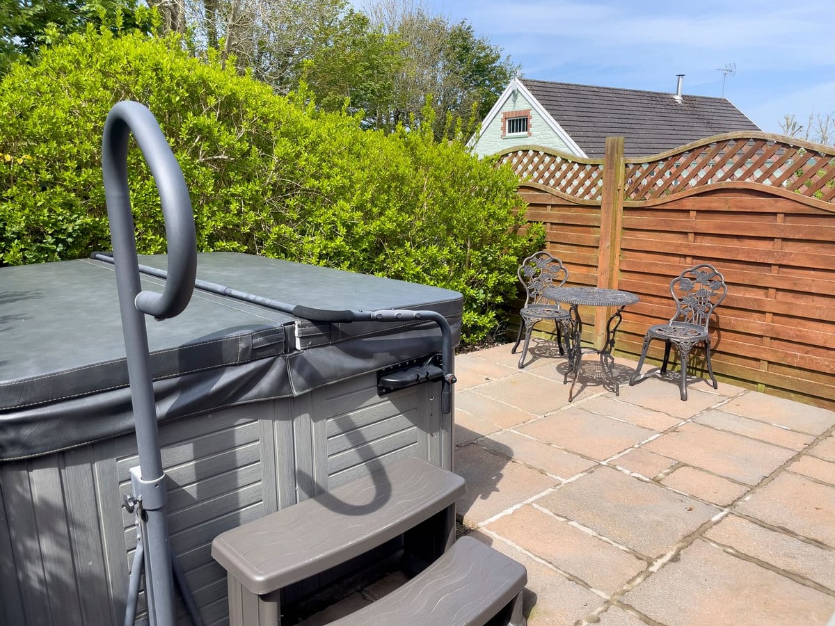 Pen-y-Bryn paved patio with a hot tub, its lid on, a bistro table and chairs, hedge and wooden fence
